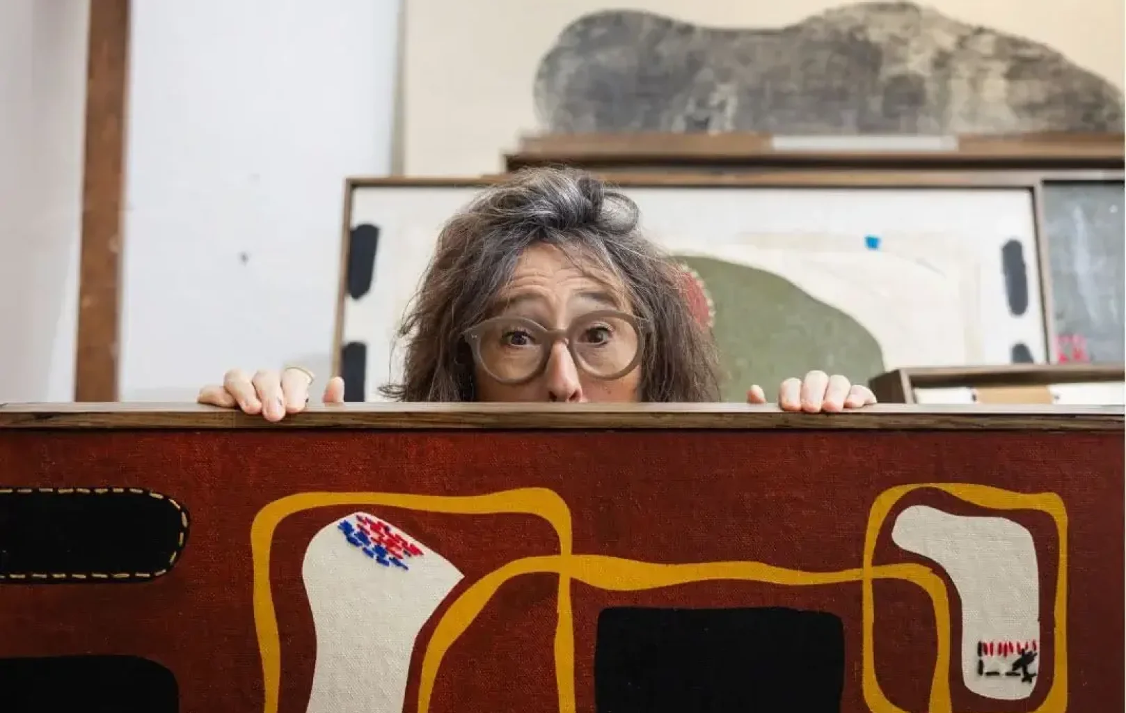 A person with glasses peeks over the top of an abstract painting in an art studio, with framed artwork and canvases in the background.