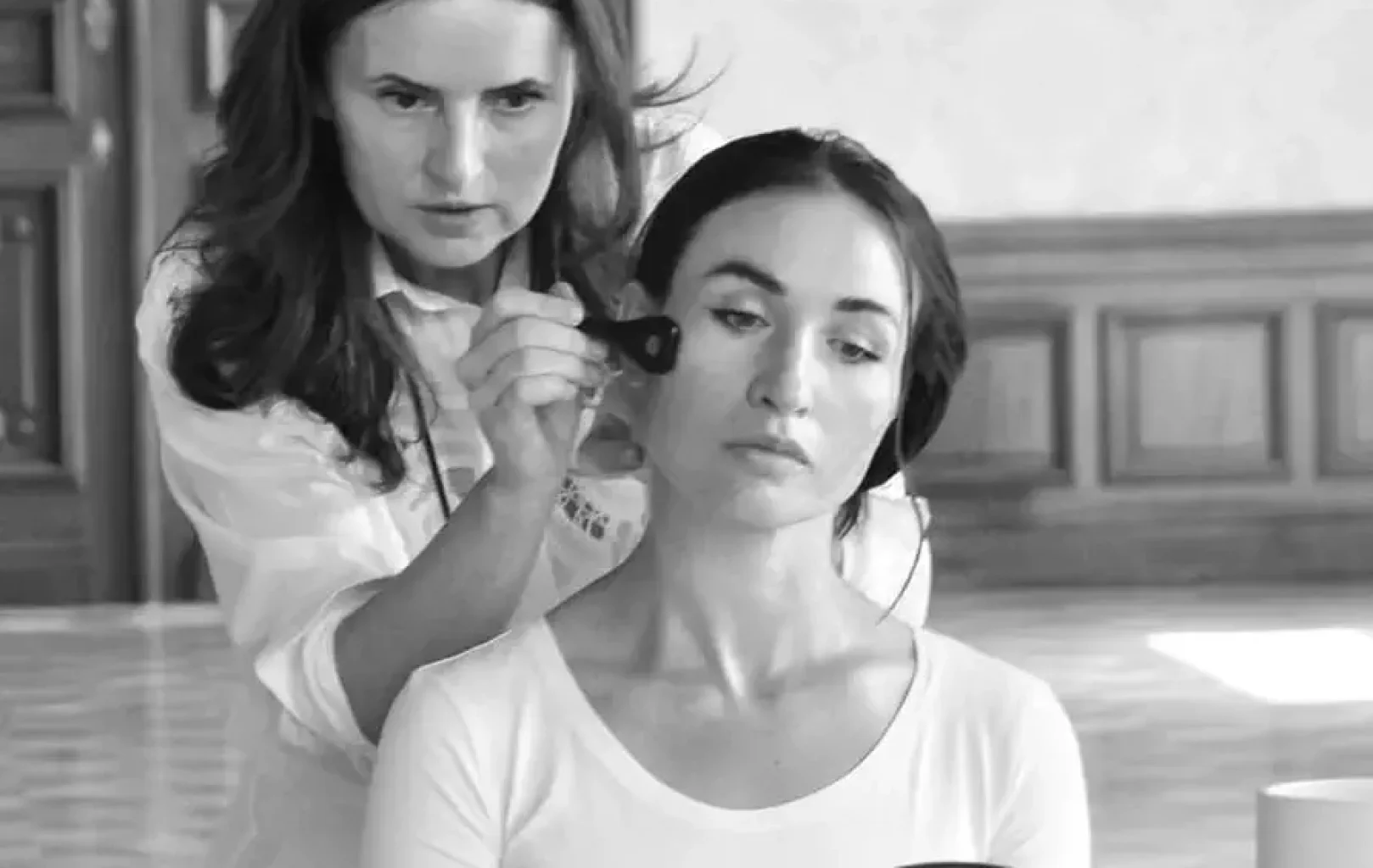 A woman from Me Myself Vienna applies makeup to another woman seated in front of a mirror in an elegant room with wooden paneling.