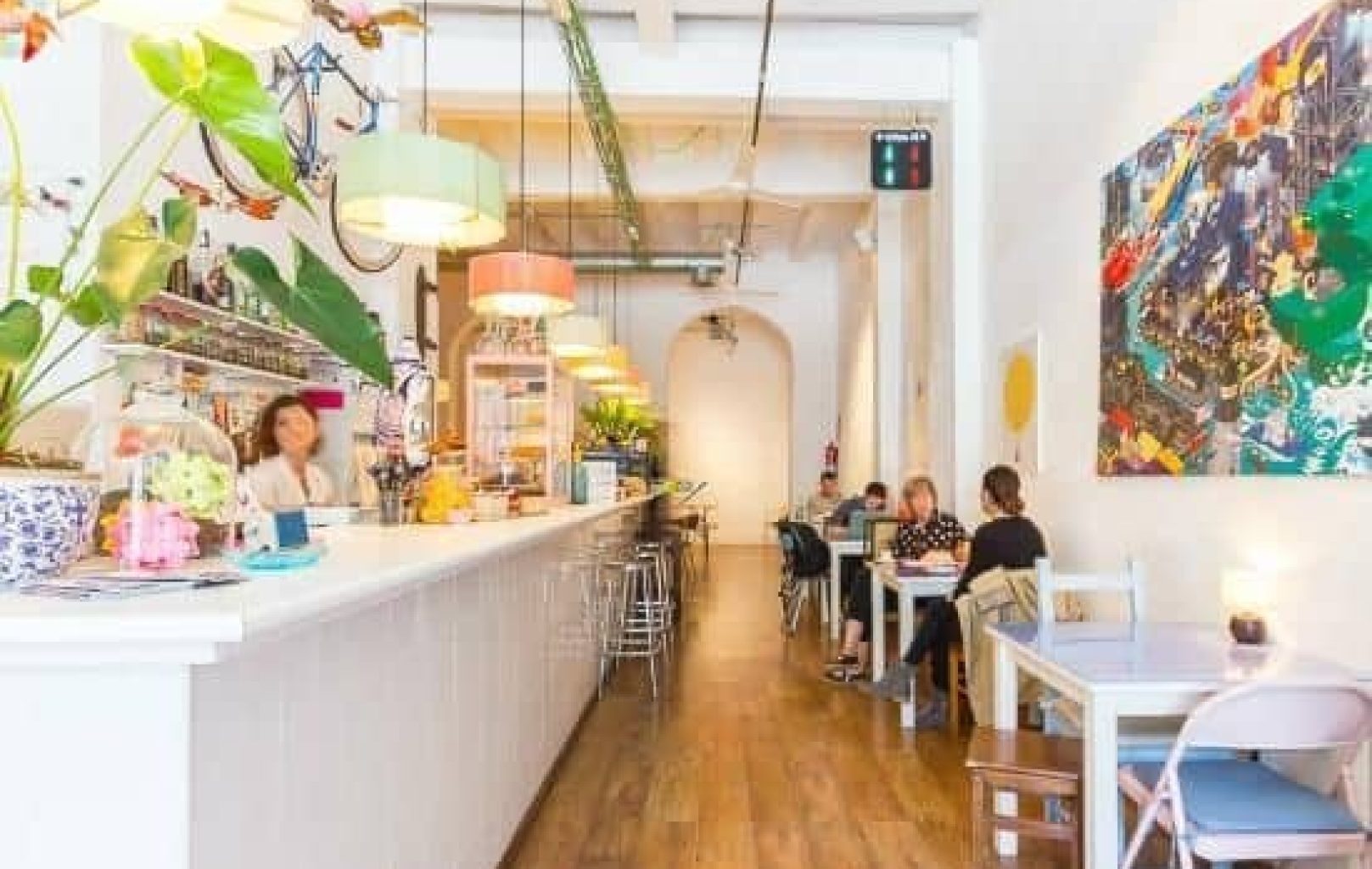 Luminosa y moderna cafetería con paredes blancas, lámparas colgantes de colores, un largo mostrador, taburetes de bar y gente sentada en las mesas laterales.