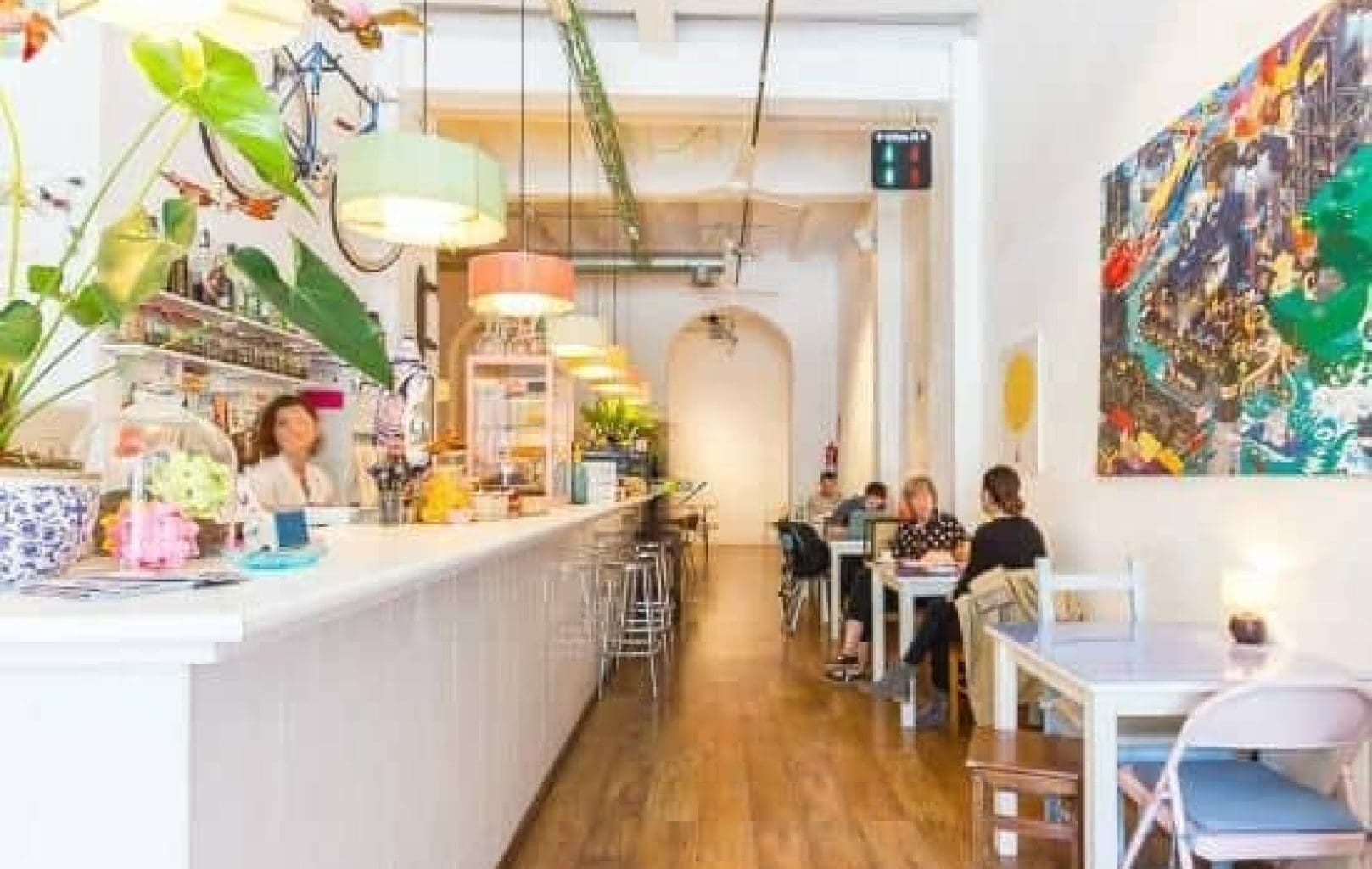 Bright, modern café with a long white counter, colorful hanging lights, and several people seated at tables along the wooden floor.