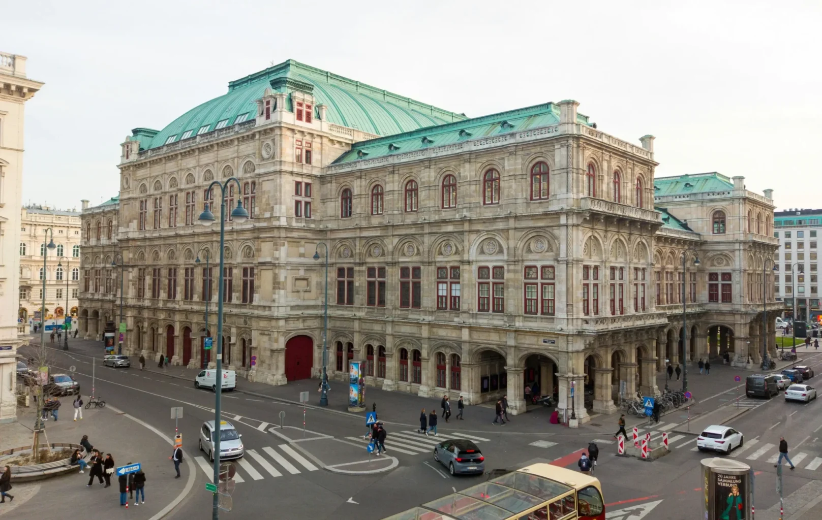 A large historic building, St. Stephens Cathedral, with a green roof and ornate stone facade sits at a busy city intersection with cars, pedestrians, and a tour bus nearby.