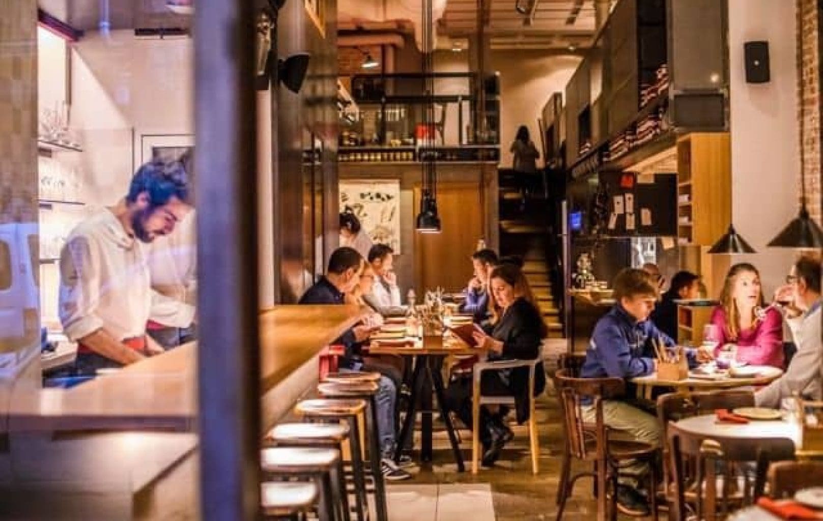 Un chef prepara la comida detrás de un mostrador mientras los comensales se sientan en las mesas y comen en el interior de un restaurante moderno y cálidamente iluminado.