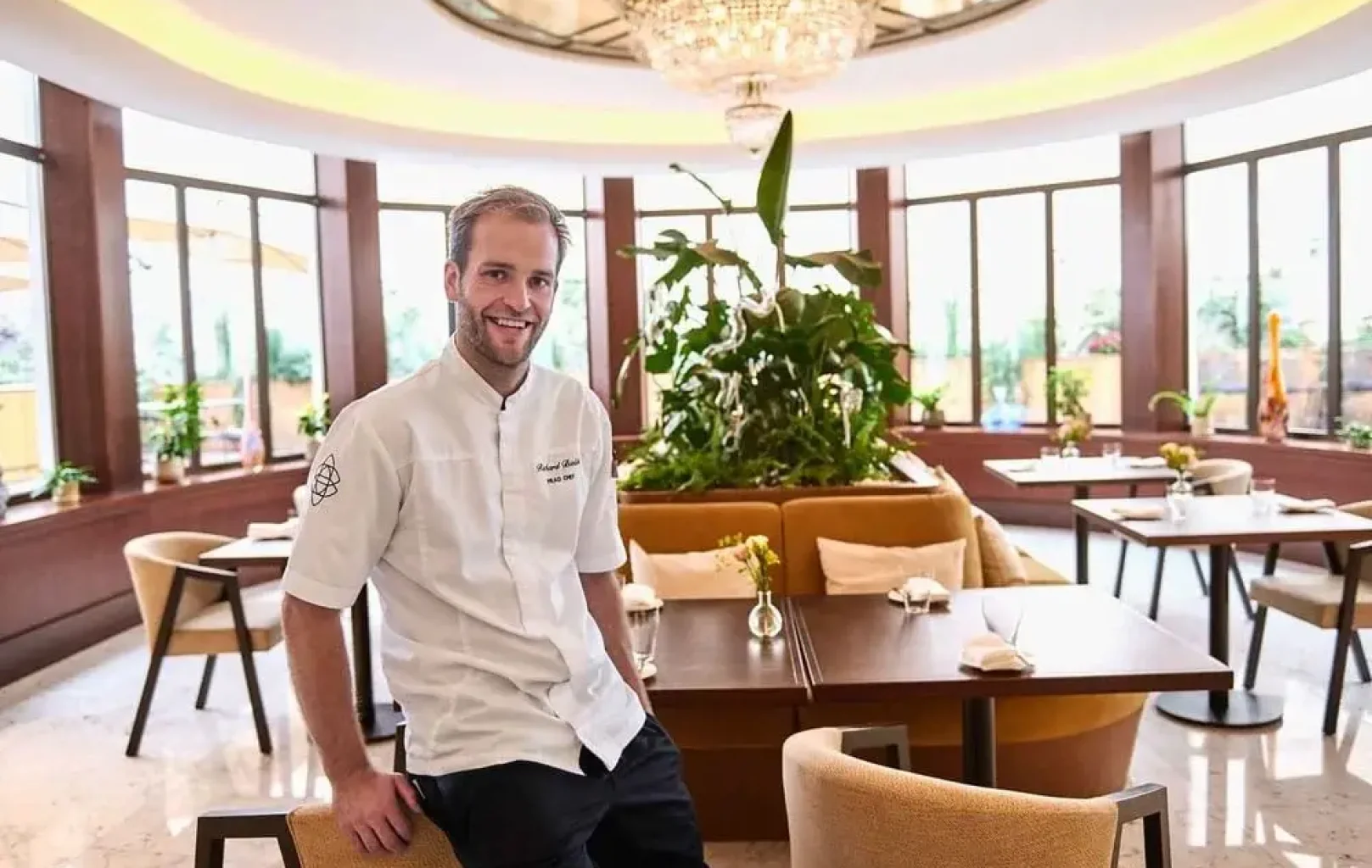 Šéfkuchař v bílé uniformě sedí na kraji stolu v elegantní moderní restauraci s velkými okny, opálenými židlemi a centrálně umístěnými rostlinami.