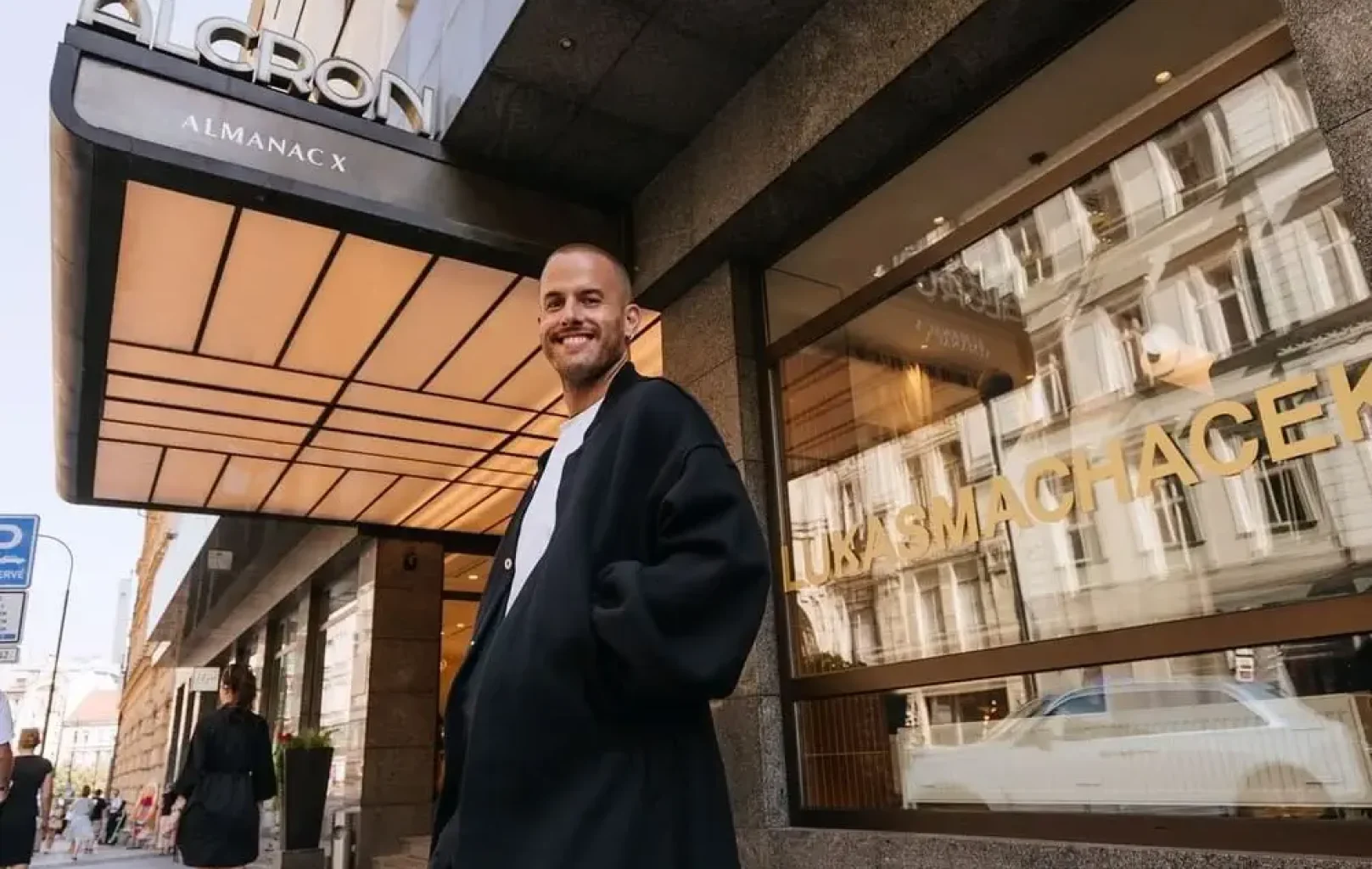 A person in a black coat stands on a sidewalk outside the Alcron Almanac X hotel, where city buildings are reflected in the window—an inviting scene for Signature Tours.