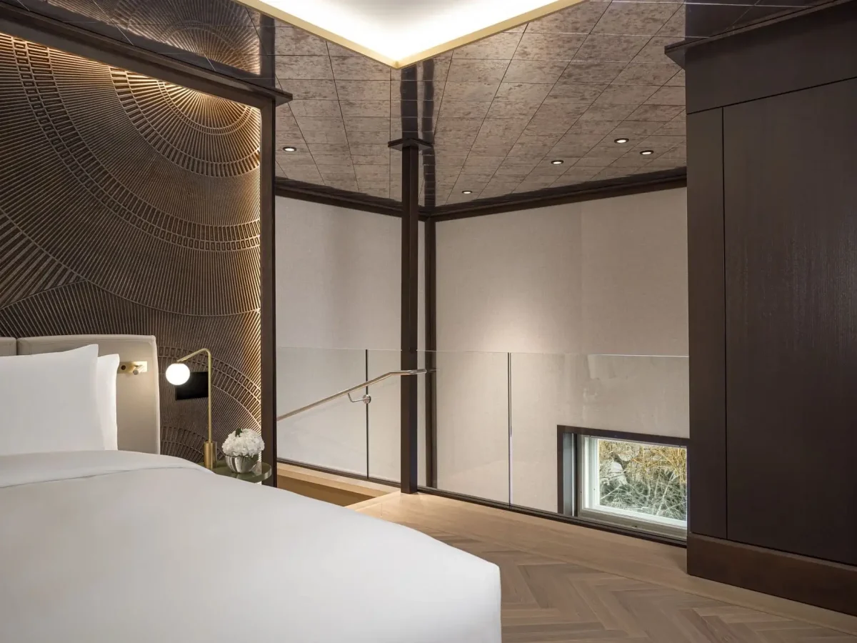 Modern duplex bedroom with partial view of a white bed, textured wall, wood paneling, glass railing, and a small window near the floor—ideal for a Park Suite retreat.