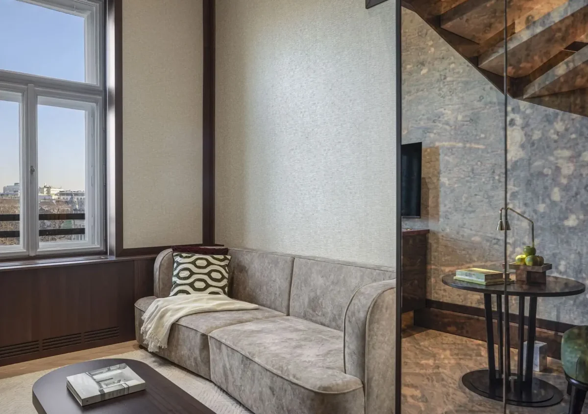 A modern Park Suite living room with a beige sofa, patterned pillow, throw blanket, coffee table with books, large window, and a glass partition revealing a small round side table in this stylish duplex.