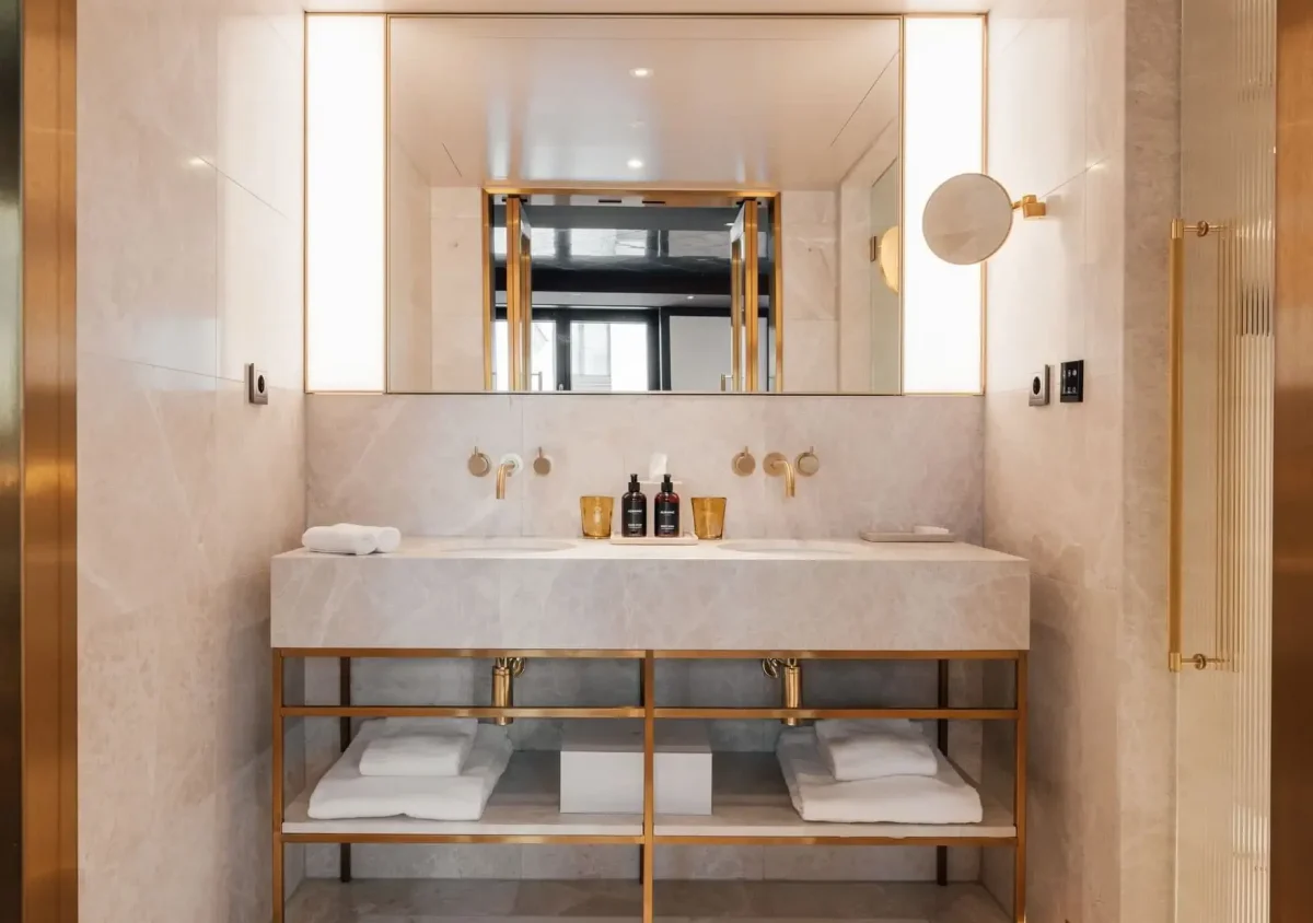 Modern bathroom in the Duplex Suite features a double sink vanity, gold fixtures, large mirror, white towels on open shelves, and marble walls and countertops.