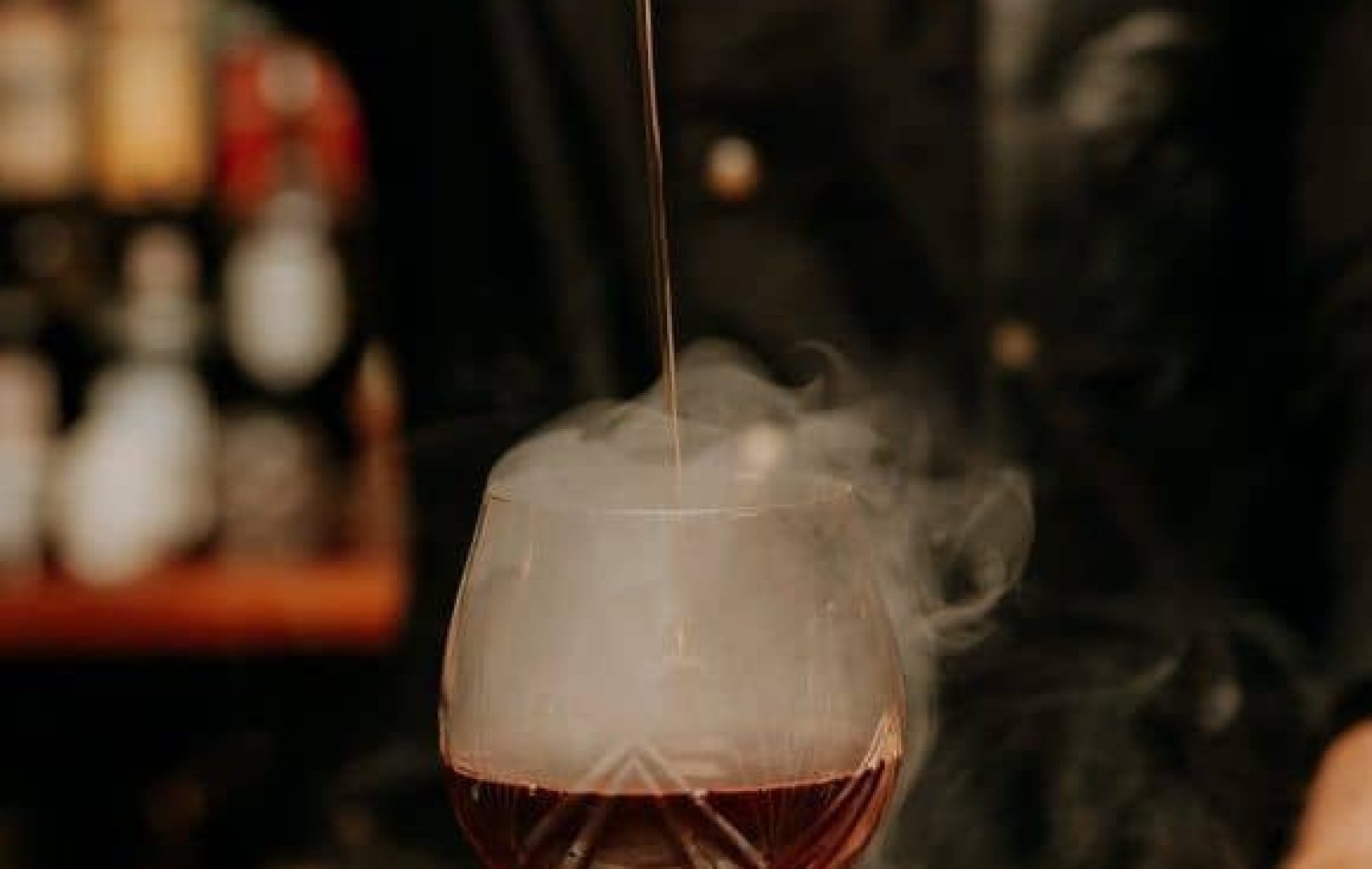 Un camarero prepara cócteles DUX en un vaso de tallo, con humo visible saliendo de la bebida, mientras vierte ginebras desde arriba.