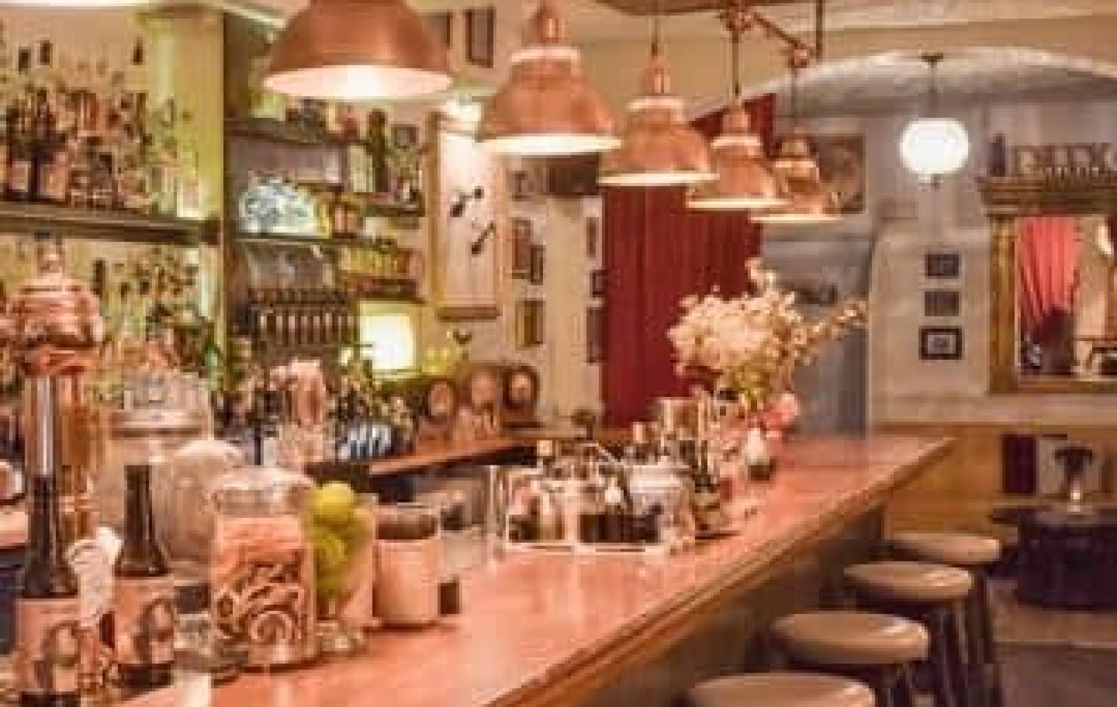 Un acogedor bar con barra de madera, taburetes, lámparas colgantes y estanterías repletas de botellas, cristalería y una tentadora selección de ginebras y cócteles.