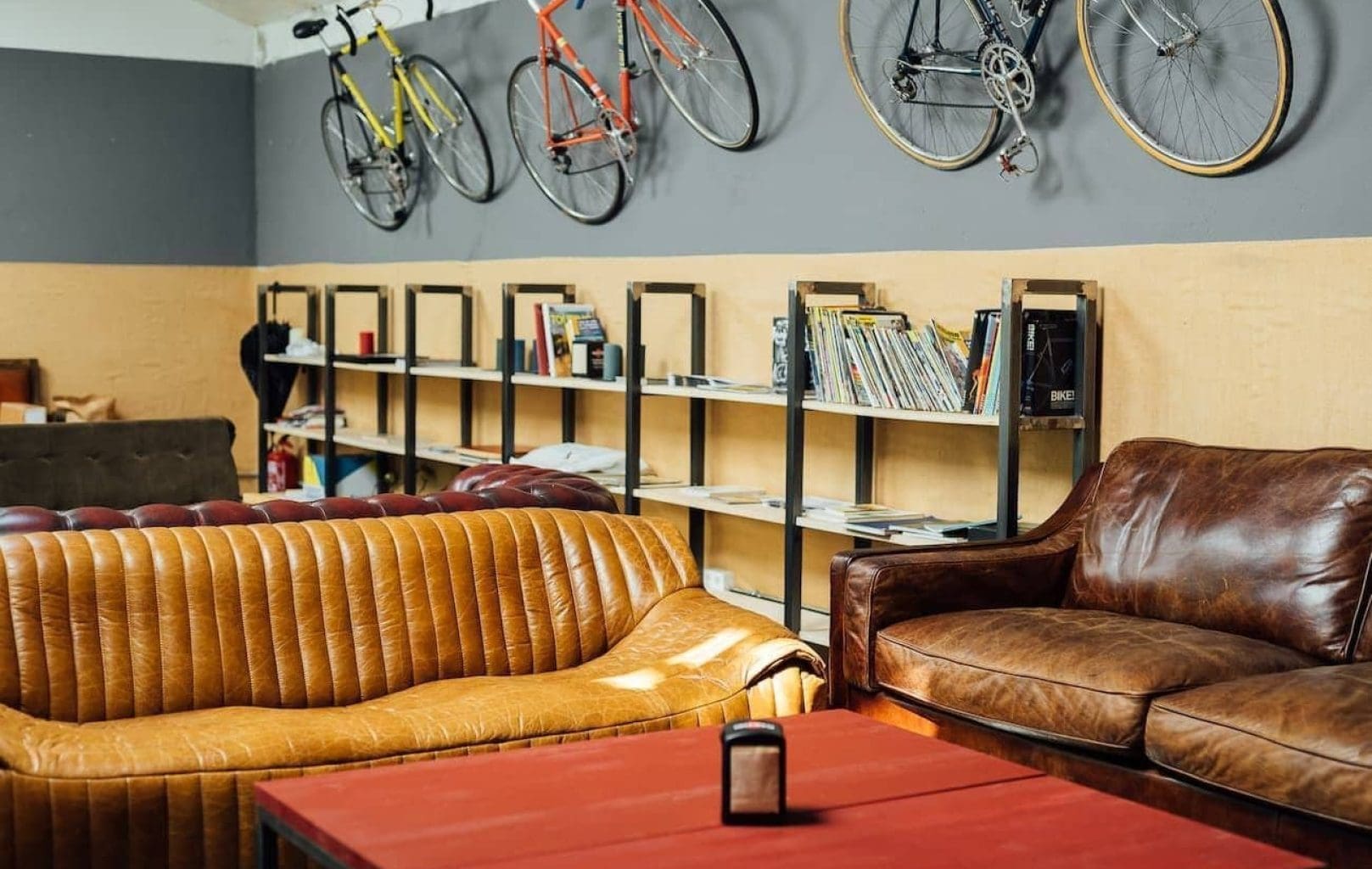 Una acogedora sala de estar con sofás de cuero marrón, una mesa de centro roja, estanterías con libros y tres bicicletas montadas en la pared.