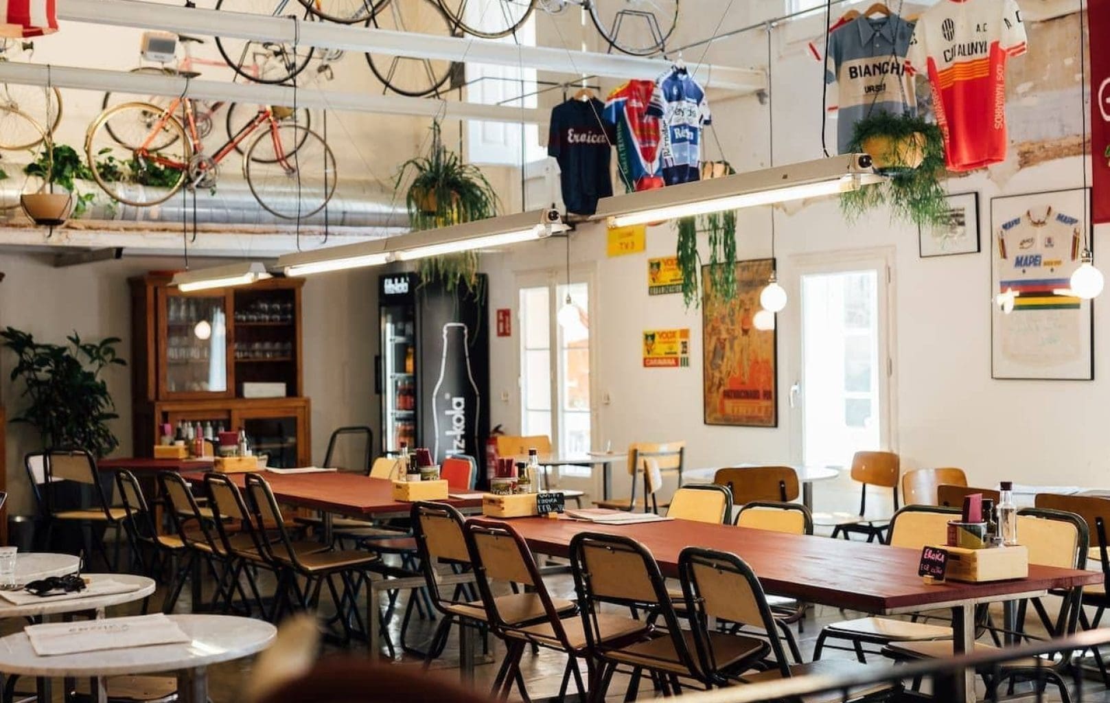 Un café con largas mesas de madera, sillas de época, bicicletas colgadas del techo y maillots de ciclismo expuestos en las paredes. La luz natural entra por grandes ventanales.