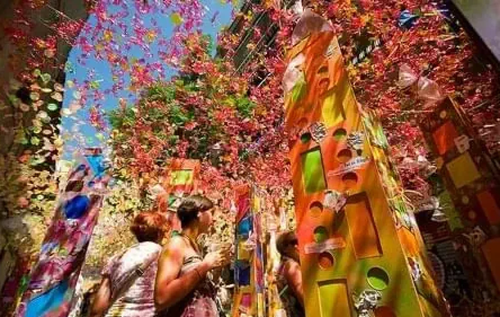La gent camina sota decoracions de colors i ornaments penjants durant un vibrant festival de carrer a l'aire lliure en un assolellat dia d'Agost.