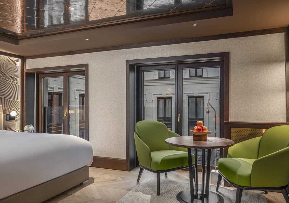 Modernes Almanac-Hotelzimmer mit einem Bett, zwei grünen Sesseln, einem kleinen runden Tisch mit Obst, großen Fenstern und neutralem Dekor.