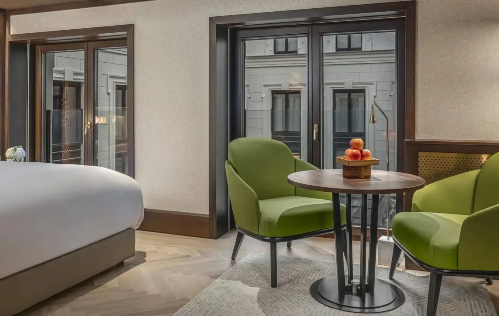 The Almanac Room features a cozy bed, two green chairs, a round table with a fruit basket, and large windows overlooking another building.