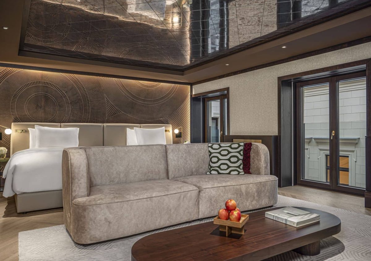 Modern Junior Suite featuring a large bed, beige sofa, low wooden coffee table with apples and a book, and ceiling mirrors reflecting the spacious interior.