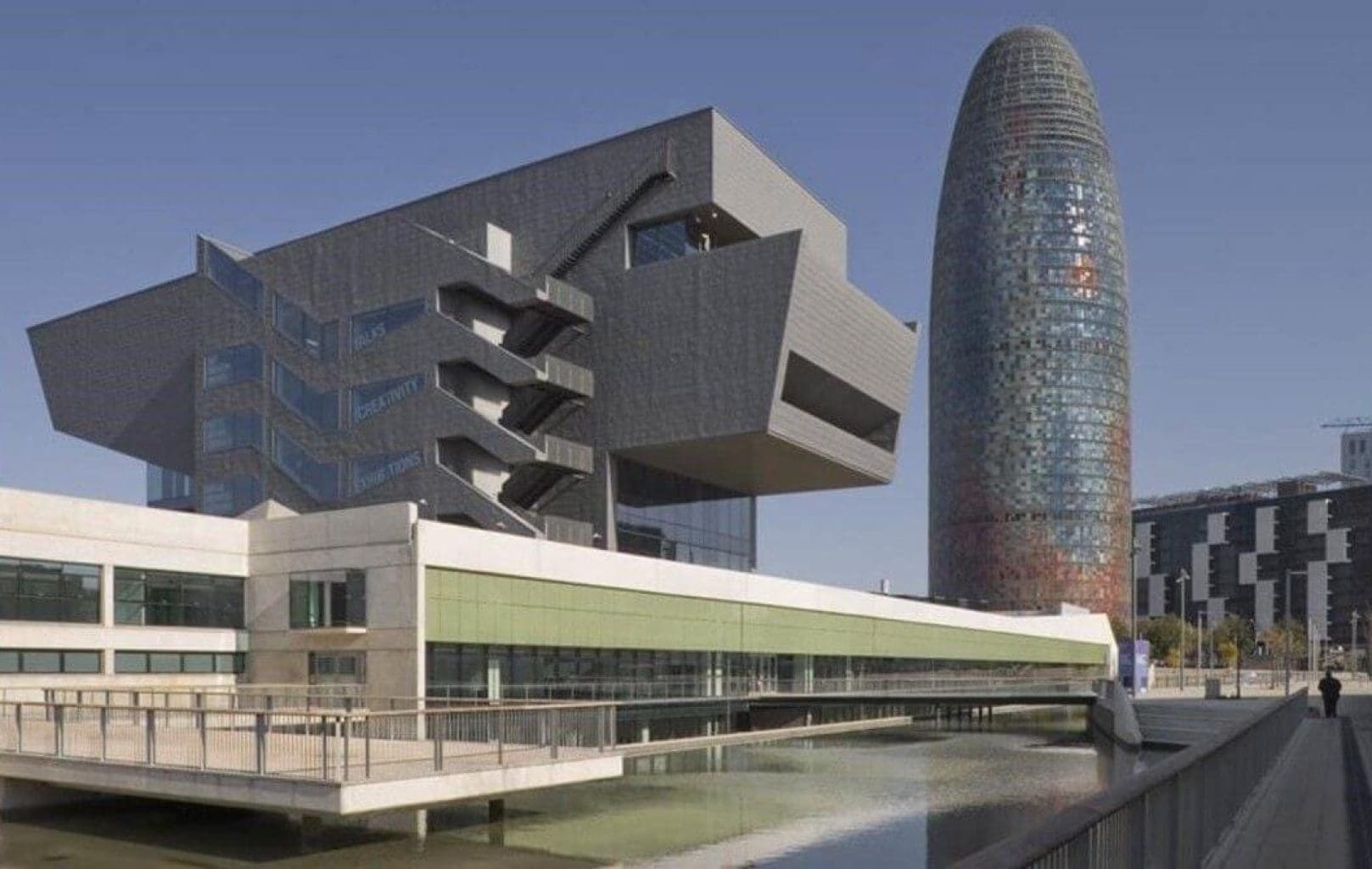 Modern architectural buildings with geometric shapes beside a reflective pool, featuring the cylindrical Torre Glòries in Barcelona under a clear sky, evoke the spirit of street art and creativity seen in Banksy’s works.