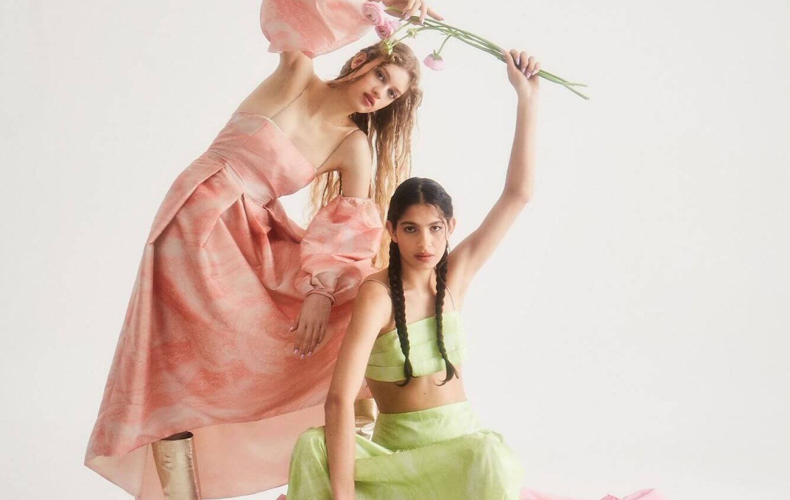 Dos mujeres posan con atuendos en tonos pastel en el Castillo de Montjuïc; una lleva un vestido rosa con mangas abullonadas que sostiene flores, mientras que la otra está sentada con un top y una falda verde claro sobre una tela rosa drapeada en medio del ambiente de la fortaleza.