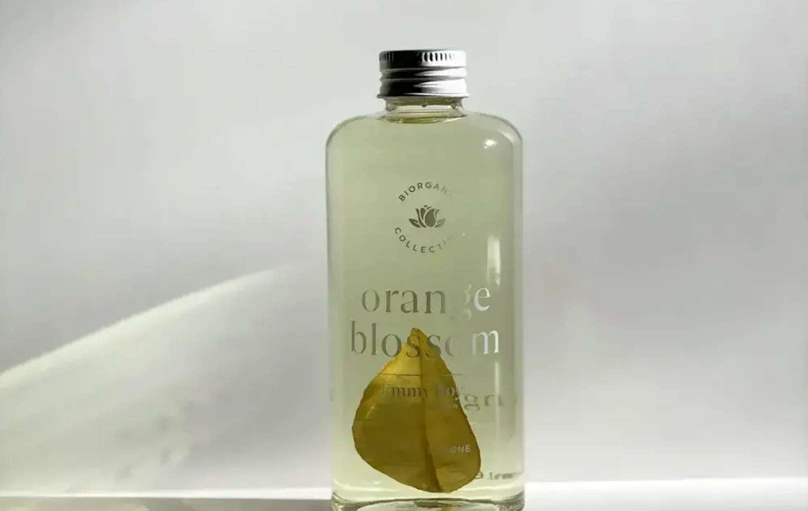 Una ampolla transparent etiquetada com a "flor de taronger" de Jimmy Boyd fragàncies, amb un tap metàl·lic, conté un líquid transparent i una sola fulla a l'interior, sobre un fons blanc llis.