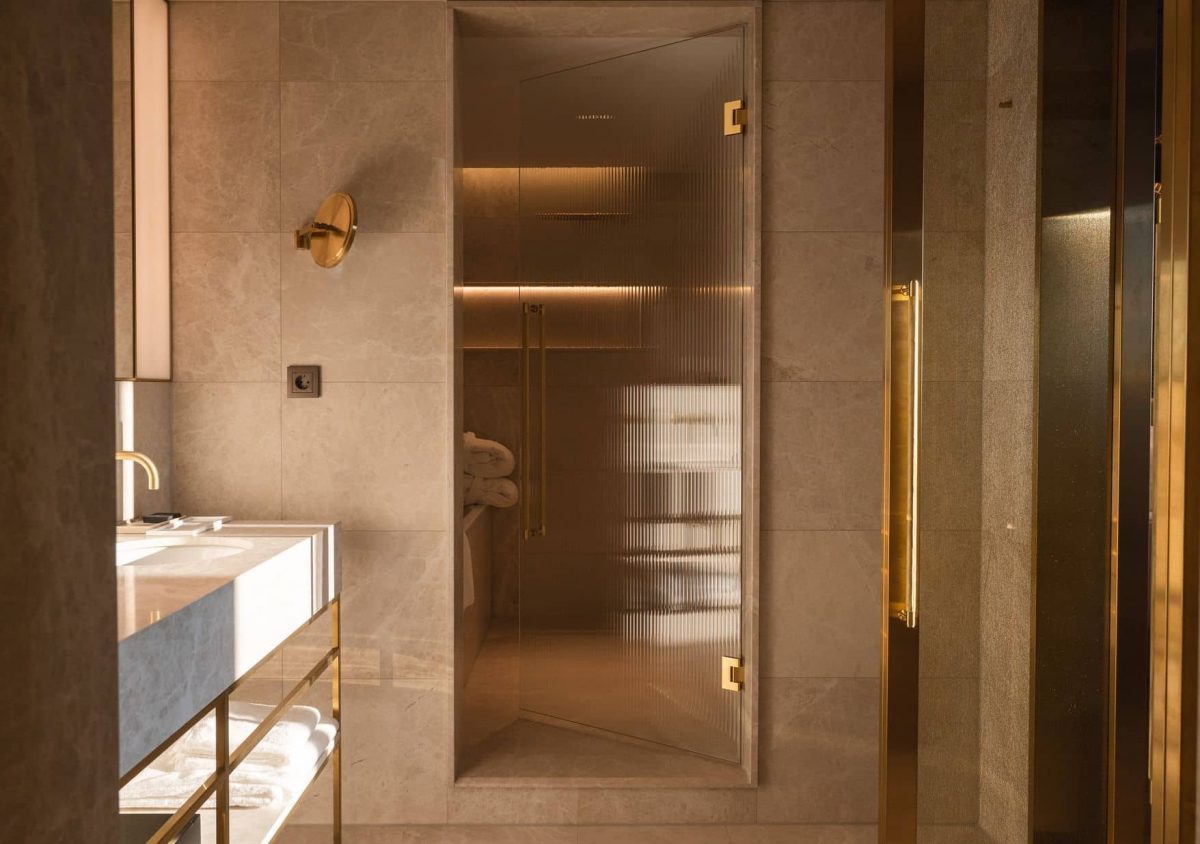 A modern Junior Suite bathroom features a glass shower door, gold fixtures, a marble sink countertop, and beige tiled walls, all illuminated by warm lighting.