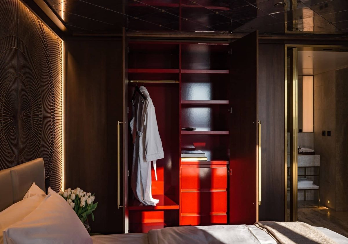 Ein modernes Schlafzimmer mit einem offenen Kleiderschrank mit Bademantel, Regalen und Schubladen, einem Bett mit weißer Bettwäsche und Tulpen auf dem Nachttisch und einem Blick in ein Badezimmer.