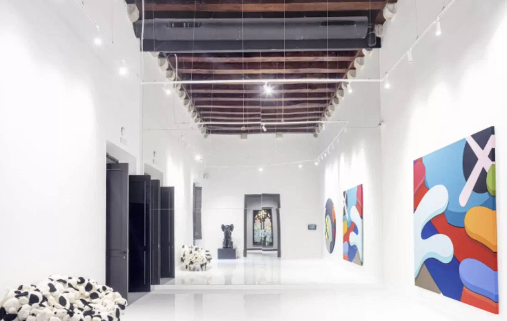 Una galería de arte moderno, como el Museo Moco de Barcelona, con paredes blancas, techo de vigas de madera, cuadros abstractos de colores, un banco manchado y obras de arte escultóricas al fondo.