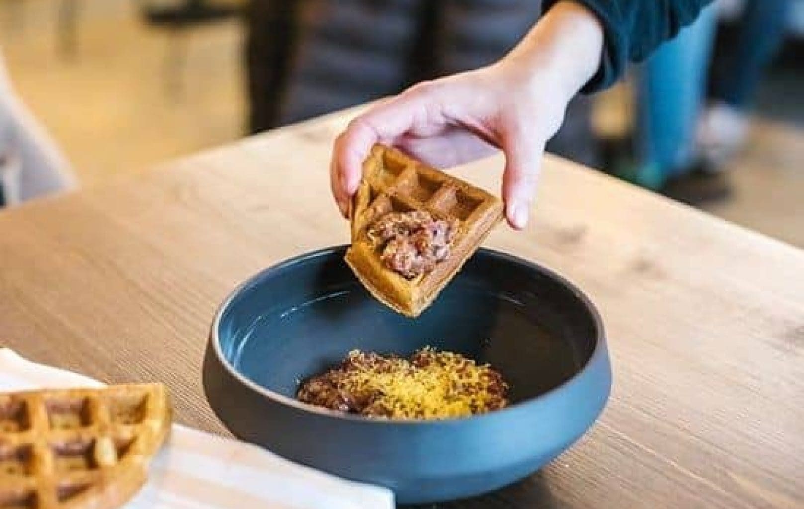 A hand dips a piece of waffle with a topping into a black bowl filled with a brown mixture and grated yellow garnish on a wooden table at La Tartareria.