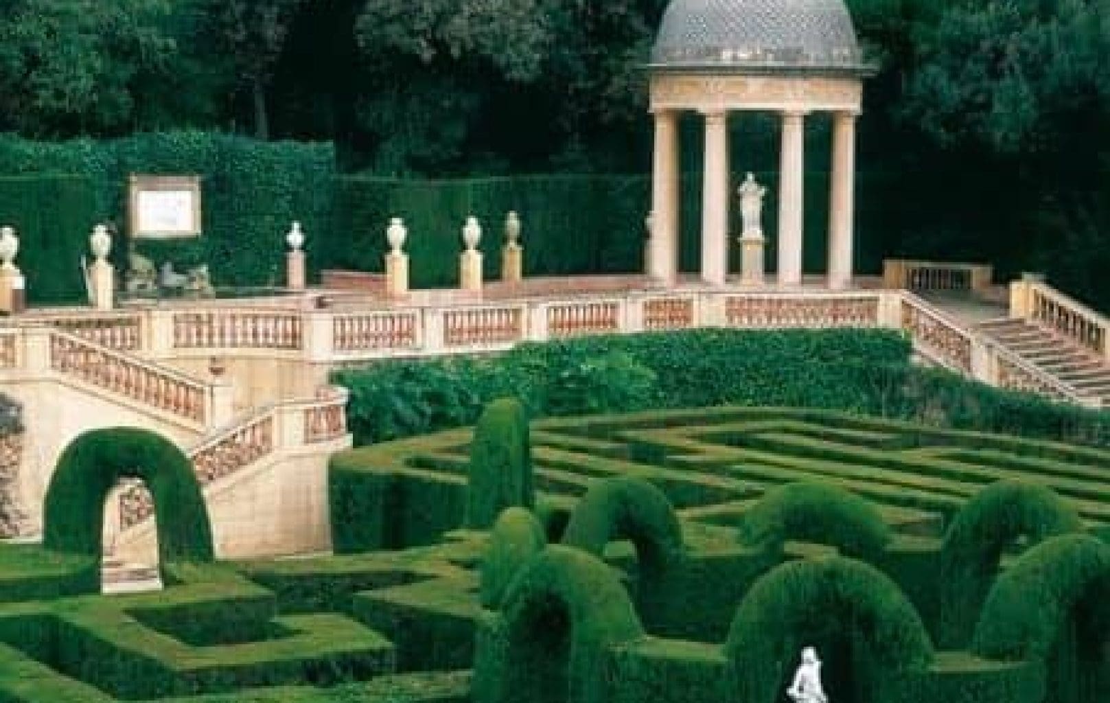 Un jardí formal del PARC LABERINT D'HORTA de Barcelona compta amb un laberint de bardisses, estàtues de marbre, un pavelló amb cúpula, una escala ornamentada i una vegetació cuidada envoltada d'arbres.