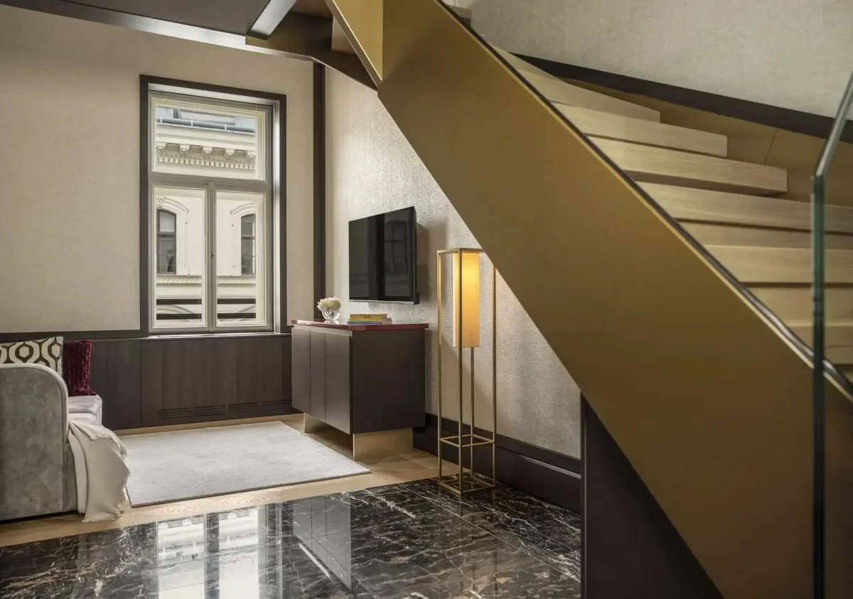 Modernes Wohnzimmer der Duplex-Suite mit Marmorboden, goldener Treppe, Wandfernseher, Schrank, Stehlampe, Sofa und großem Fenster mit Blick auf ein klassisches Gebäude.