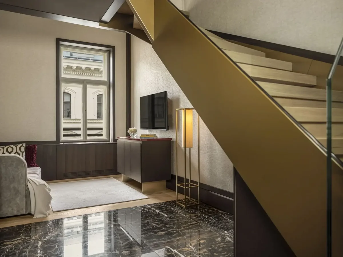 Modernes Wohnzimmer der Duplex-Suite mit Marmorboden, goldener Treppe, Wandfernseher, Schrank, Stehlampe, Sofa und großem Fenster mit Blick auf ein klassisches Gebäude.