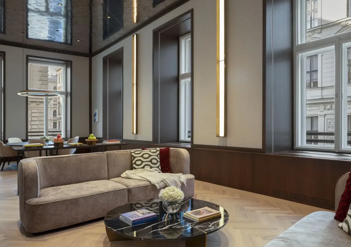 Moderner Lounge-Bereich mit beigen Sofas, einem schwarzen Marmor-Couchtisch, Büchern und großen Fenstern; kunstvoll gestaltet im Stil einer Wiener Suite, mit einem Essbereich im Hintergrund.