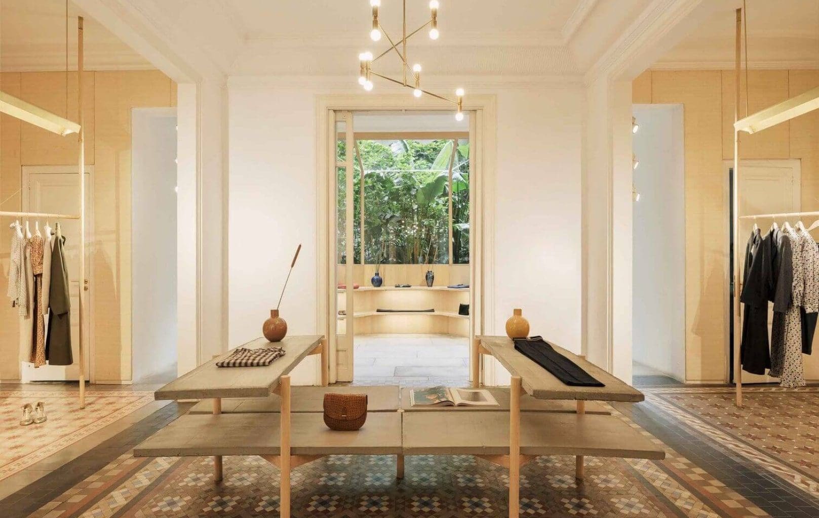 Botiga minimalista amb terra de rajoles estampades, penjadors de roba a banda i banda, dues taules expositores al centre i grans finestrals a la part posterior que ofereixen vistes que recorden la vegetació que envolta la famosa fortalesa de Barcelona, el Castell de Montjuïc.