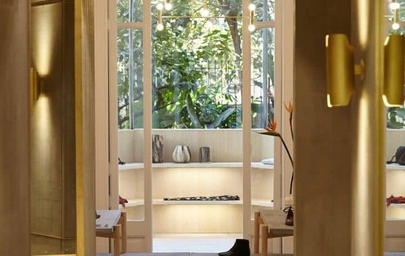 Un interior de boutique moderno y bien iluminado, con zapatos y bolsos expuestos en estanterías, grandes ventanales y vegetación visible en el exterior.