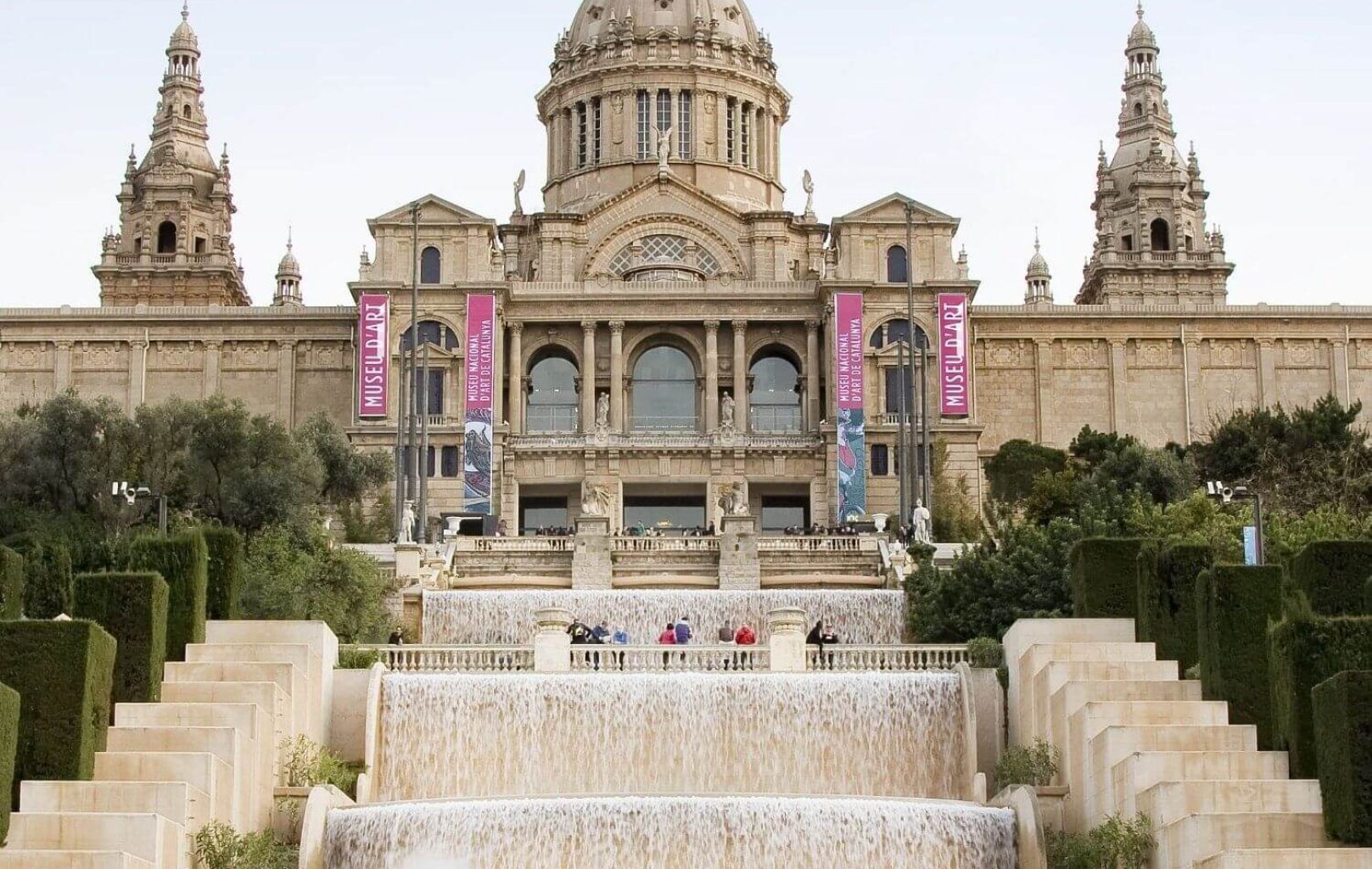 Un gran edifici històric, seu del Museu Nacional d'Art de Catalunya (MNAC), es troba darrere de cascades i fonts esglaonades, amb gent caminant i fent fotos a les escales i terrasses.
