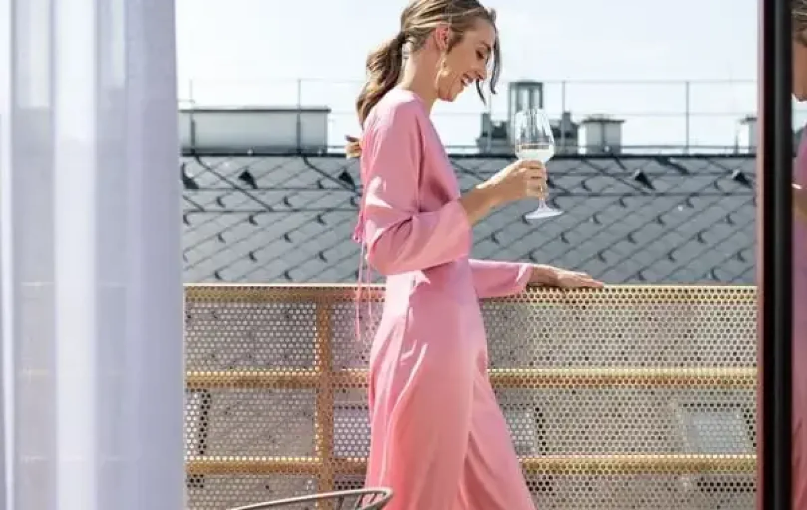 Woman in a long pink dress holding a wine glass stands on a balcony with metal railing, enjoying her summer escape in Prague, with charming rooftops visible in the background.