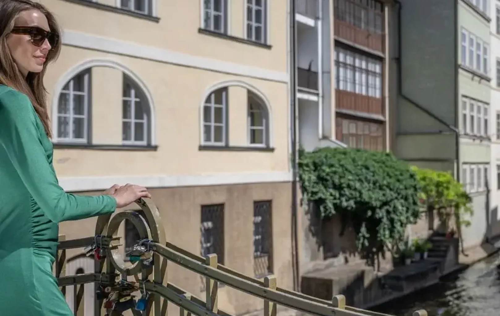 Žena v zelených šatech stojí za slunečného dne na mostě s výhledem na kanál lemovaný budovami a zachycuje podstatu prohlídek s podpisem.