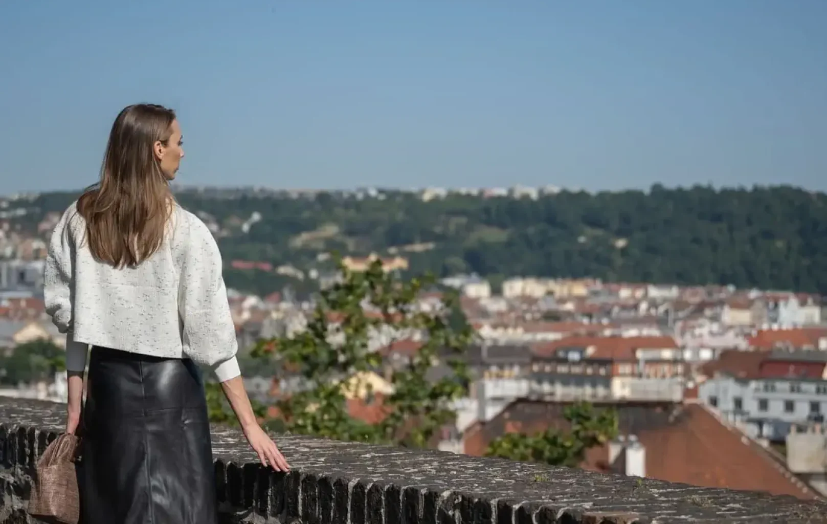 A woman in a white top and black skirt stands by a stone wall during one of our Signature Tours, overlooking a cityscape with red-roofed buildings and green hills beneath a clear blue sky.