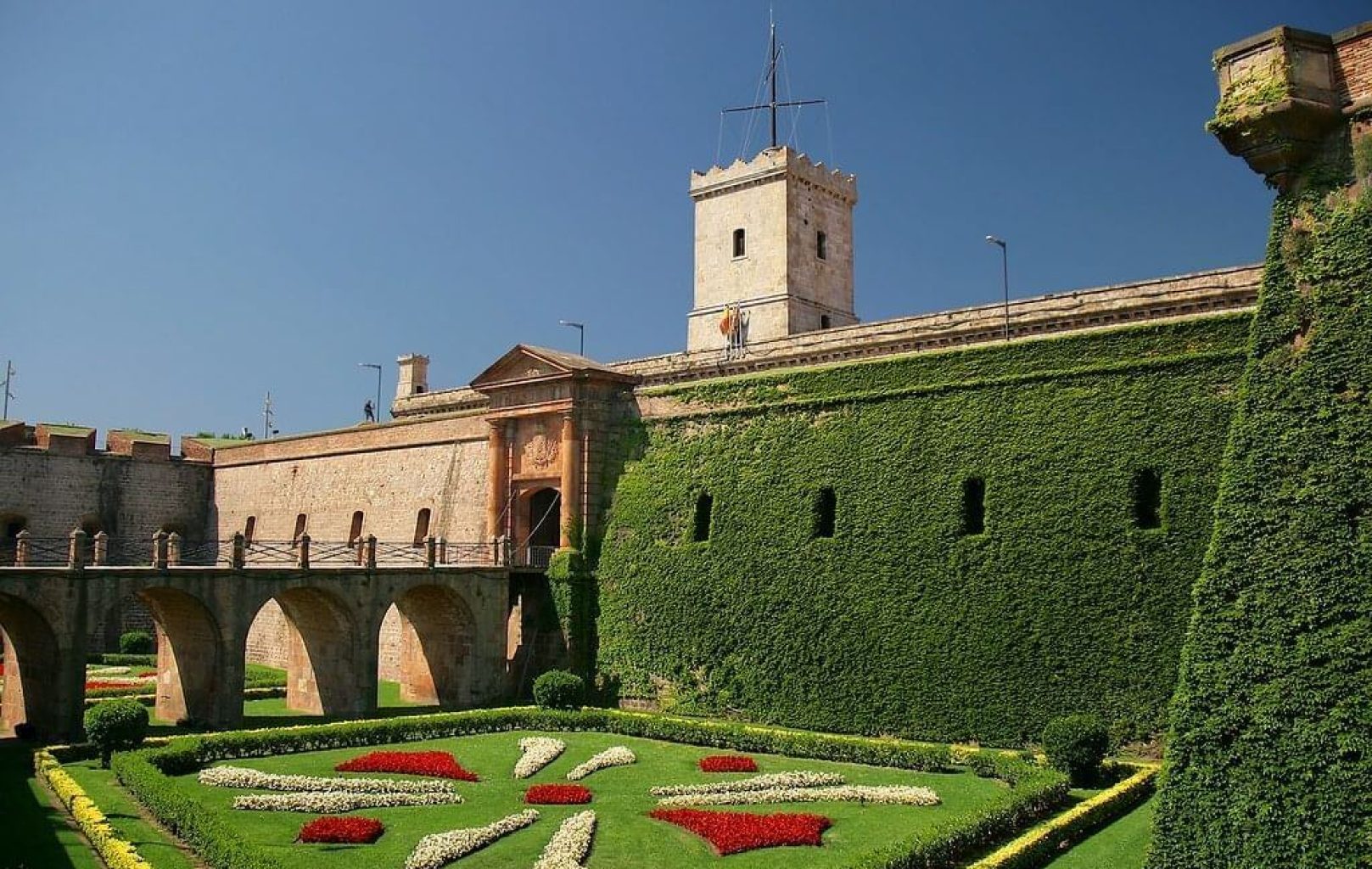 Fortaleza de piedra con una torre, un puente arqueado y muros cubiertos de hiedra -que recuerda al castillo de Montjuïc de Barcelona- con vistas a un jardín formal con setos recortados y parterres estampados bajo un cielo azul despejado.