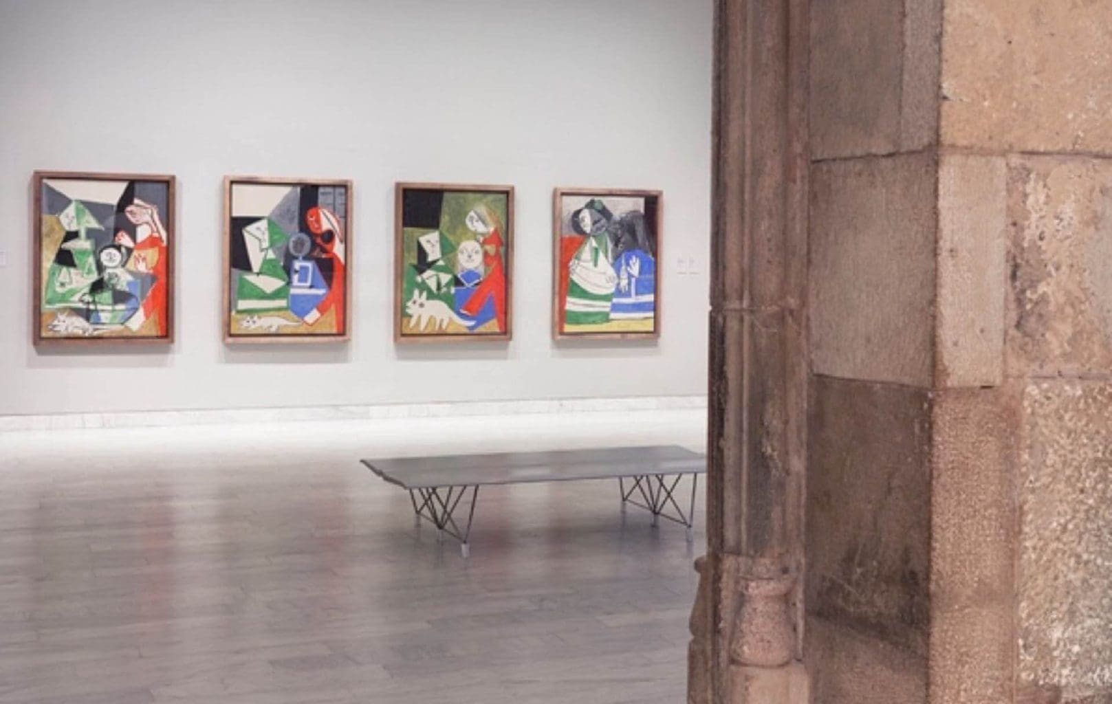 Four abstract paintings hang on a white wall in a gallery at the Picasso Museum, with a bench in front; stone columns are visible in the foreground, echoing the historic charm of Barcelona.