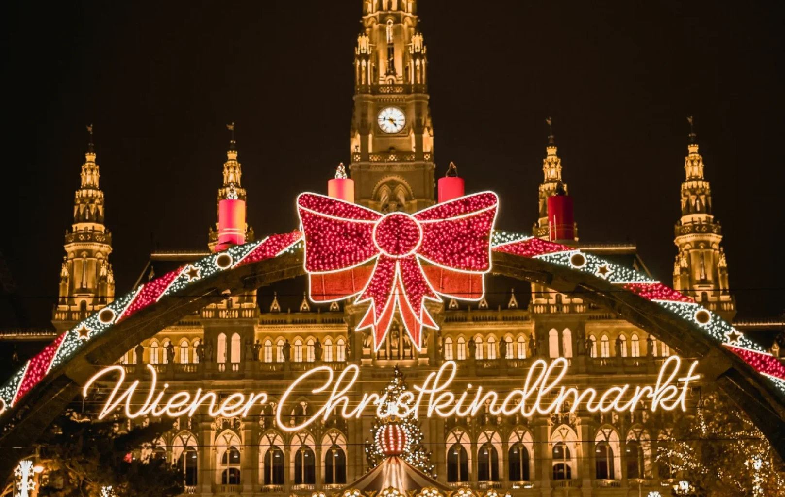 Vienna City Hall shines at night with festive lights and a large bow for the Wiener Christkindlmarkt Christmas market, while St. Stephen's Cathedral stands nearby, adding to the city's enchanting holiday ambiance.