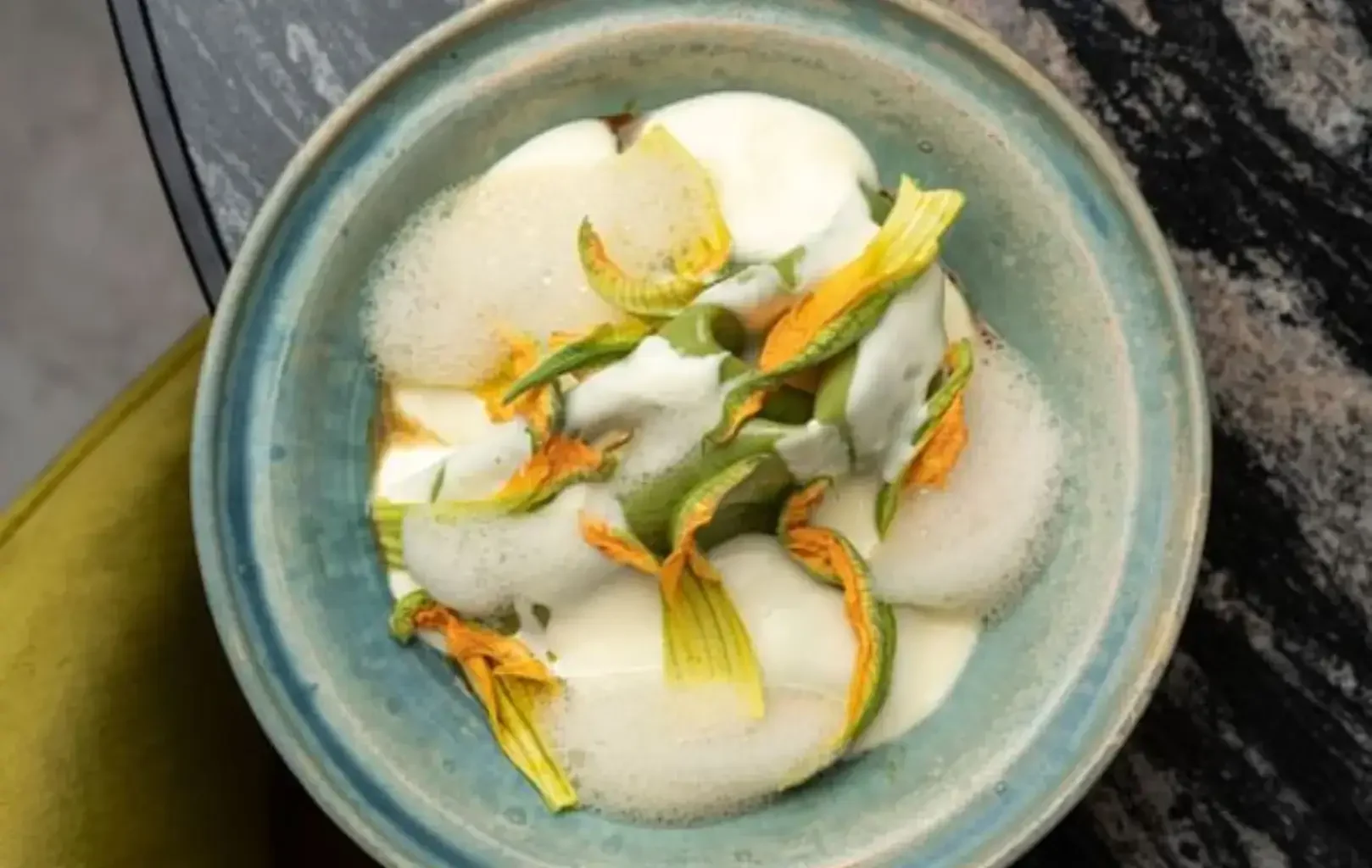 Un cuenco azul verdoso que contiene un plato cremoso, parte del Almanaque Degusta, adornado con espuma y flores amarillas de calabacín sobre una superficie oscura y jaspeada.
