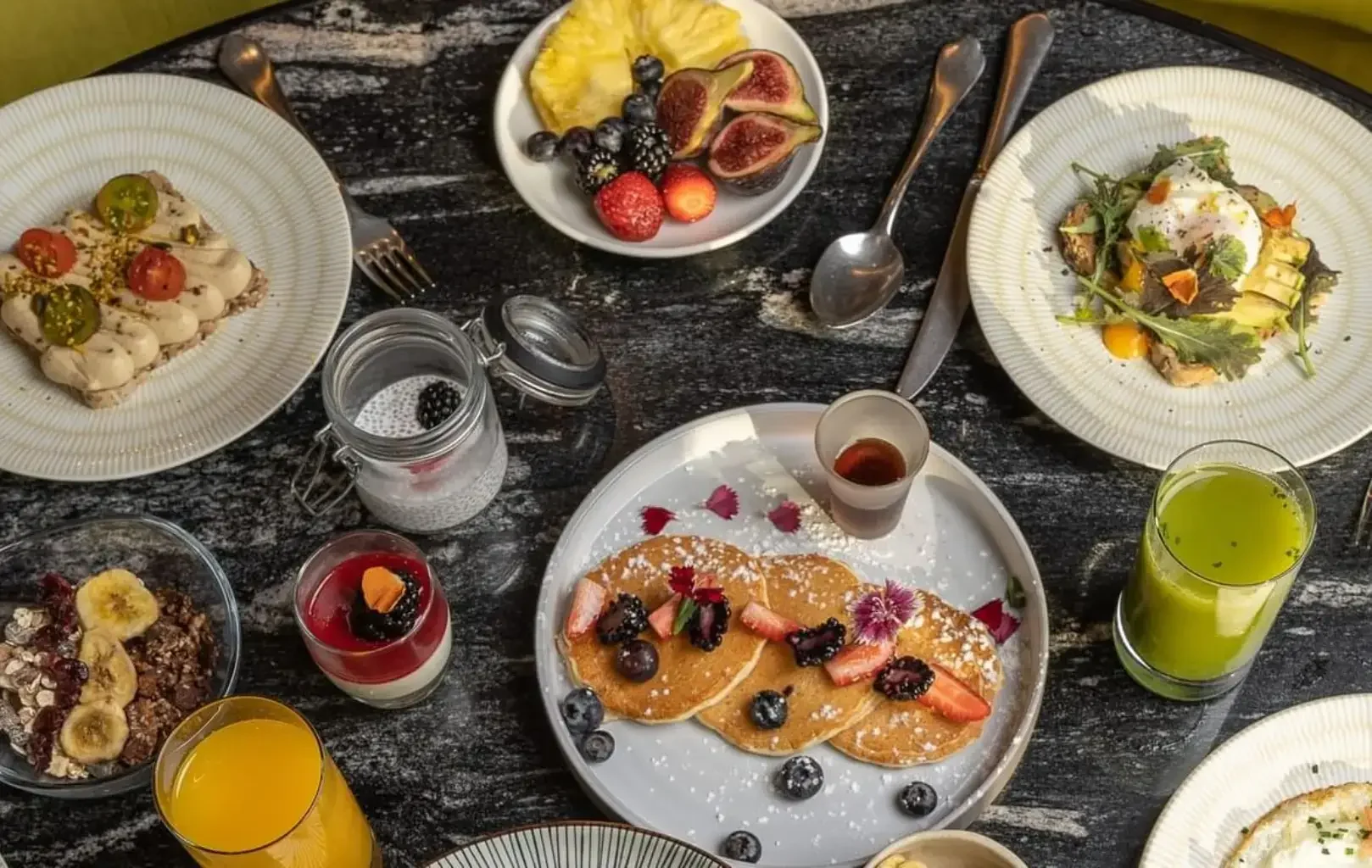 Una taula d'esmorzar parada amb creps amb fruita, torrades d'alvocat, un plat de fruita, parfaits i diversos sucs sobre una superfície de marbre fosc: una escena atractiva treta directament de les pàgines de Degusta Almanac.