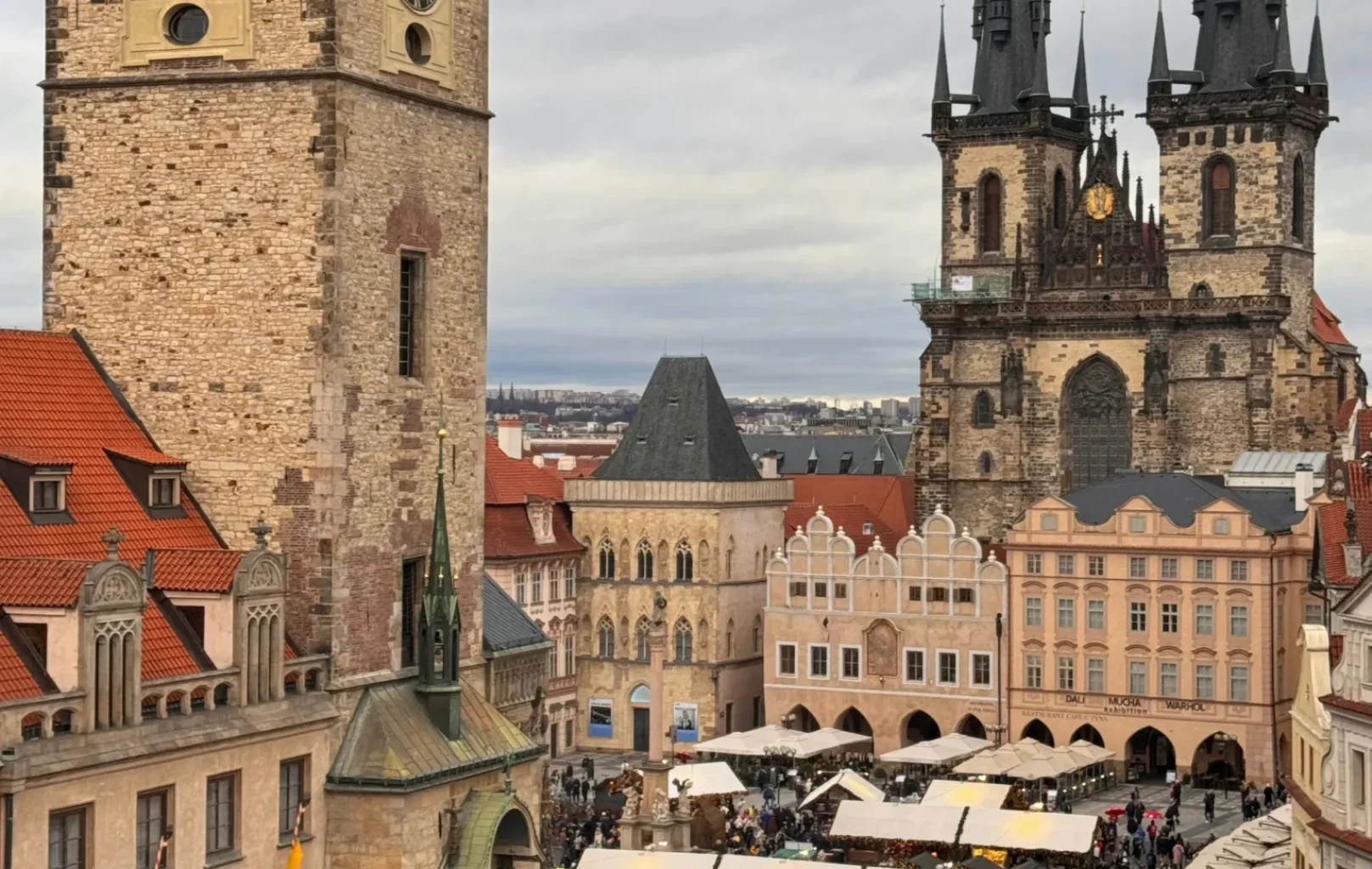 View of a busy market square in Prague, featuring the Old Town Hall with its clock tower and the Church of Our Lady before Týn in the background, not far from lively Wenceslas Square, under a cloudy sky.