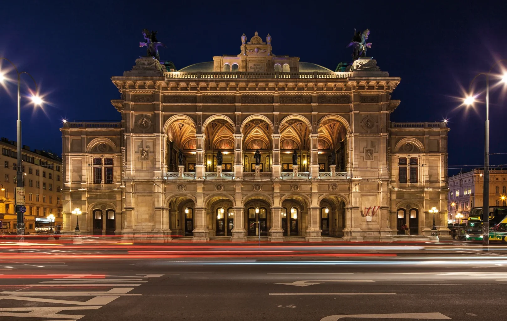 Die Wiener Staatsoper bei Nacht. Das große Opernhaus ist beleuchtet, um die beeindruckenden architektonischen Details zu enthüllen, mit Lichtspuren von vorbeifahrenden Fahrzeugen im Vordergrund.