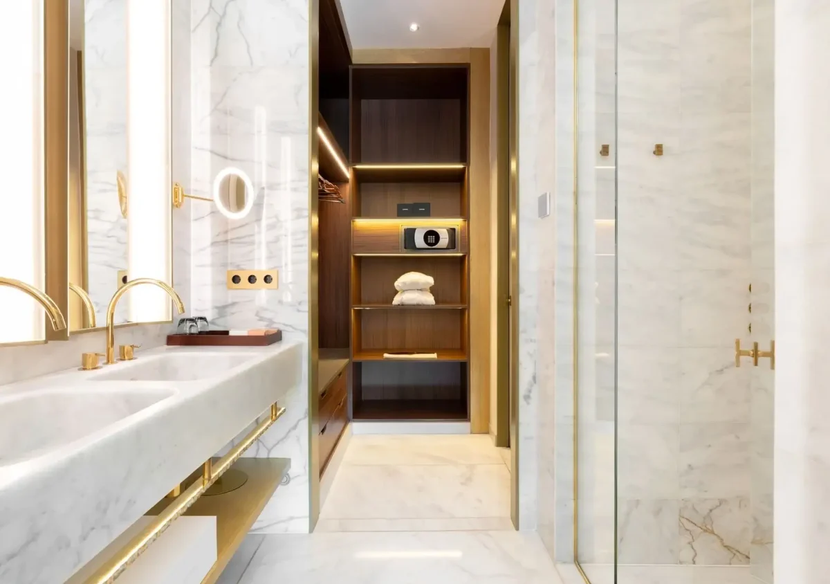 Modernes Badezimmer in der Almanac Family Suite mit Marmorwänden und -böden, Doppelwaschbecken mit goldenen Armaturen, Glasdusche sowie einem offenen Schrank mit Regalen, Handtüchern und einem Safe.