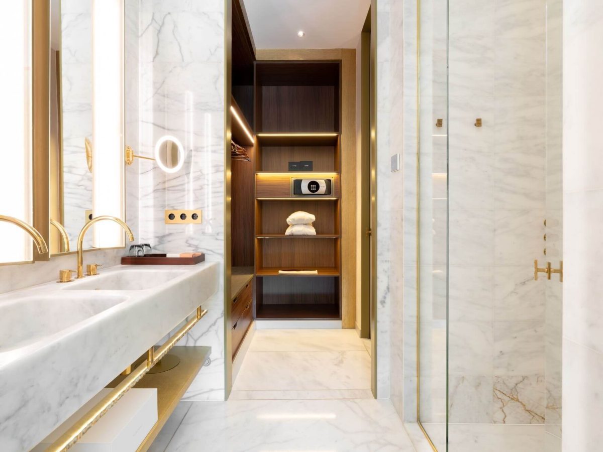 Modernes Badezimmer in der Almanac Suite mit Marmoroberflächen, Doppelwaschbecken, goldenen Armaturen, begehbarer Dusche sowie einem offenen Holzschrank mit Regalen, einem Safe und ordentlich gefalteten Handtüchern.