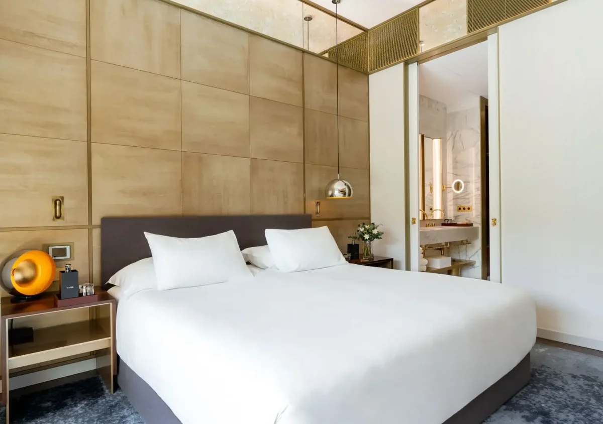 Modernes Hotelzimmer mit großem Bett, weißer Bettwäsche, holzgetäfelten Wänden, Nachttischen, modernen Lampen und einem offenen Durchgang zum Badezimmer - perfekt zum Entspannen in der einladenden Almanac Family Suite.