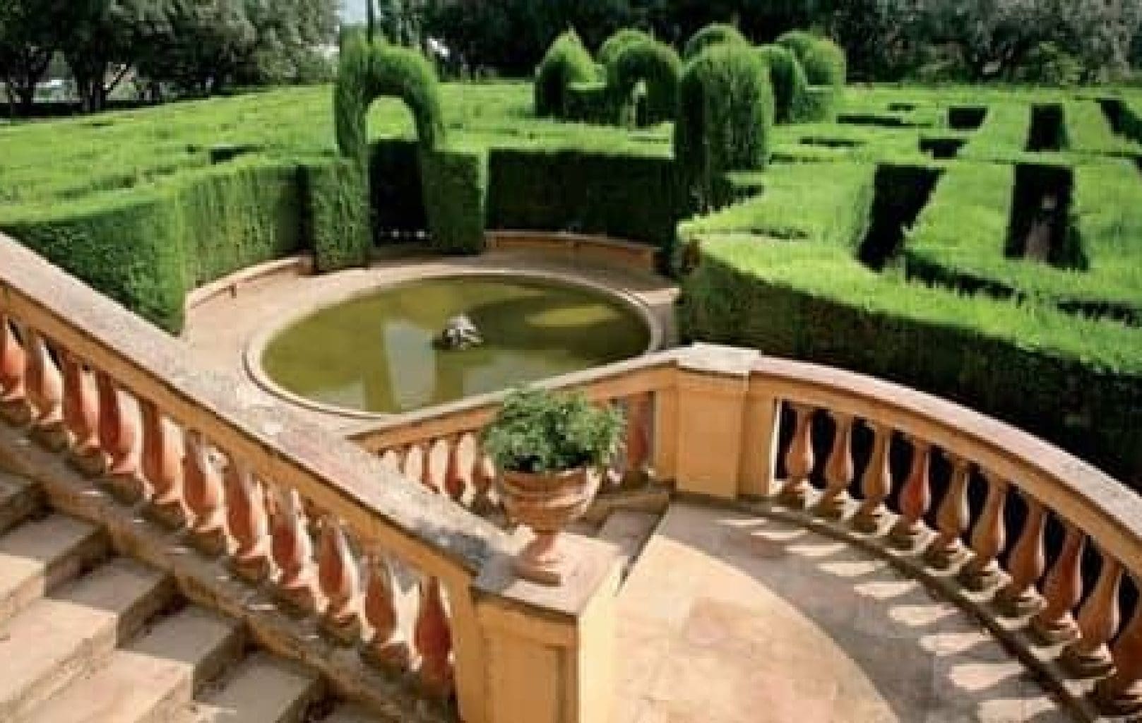Una escala de pedra amb balustrada baixa a un estany circular, envoltat de bardisses retallades que formen un laberint, que recorda el Parc Laberint d'Horta de Barcelona.