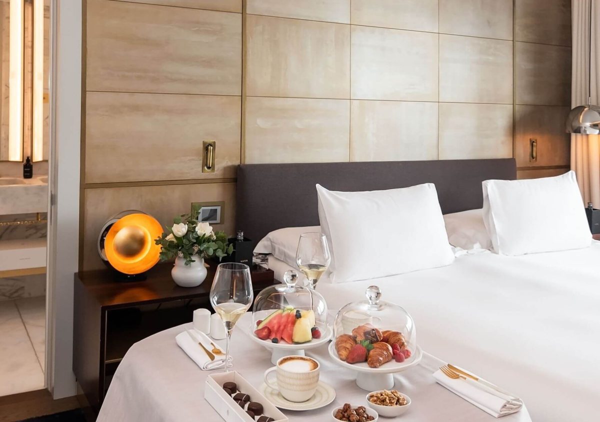 Una suite à tic amb un llit ben fet, una taula parada amb fruita, pastissos, cafè, vi i aperitius, a més d'una tauleta de nit amb una petita là mpada i flors, perfecta per a hostes que busquen luxe a les dues habitacions.
