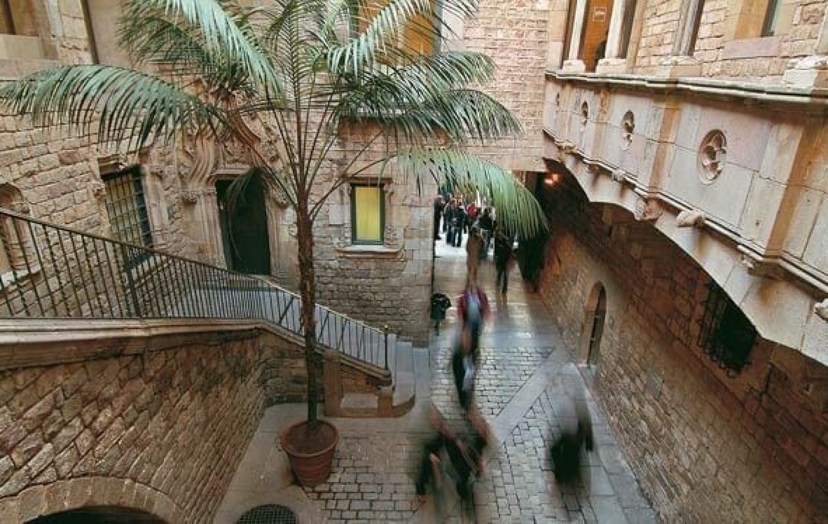 Patio de piedra del Museu Picasso con una palmera en maceta, una escalera arqueada y figuras borrosas caminando por debajo; grupos de personas se reúnen cerca de una entrada bajo balcones de piedra.