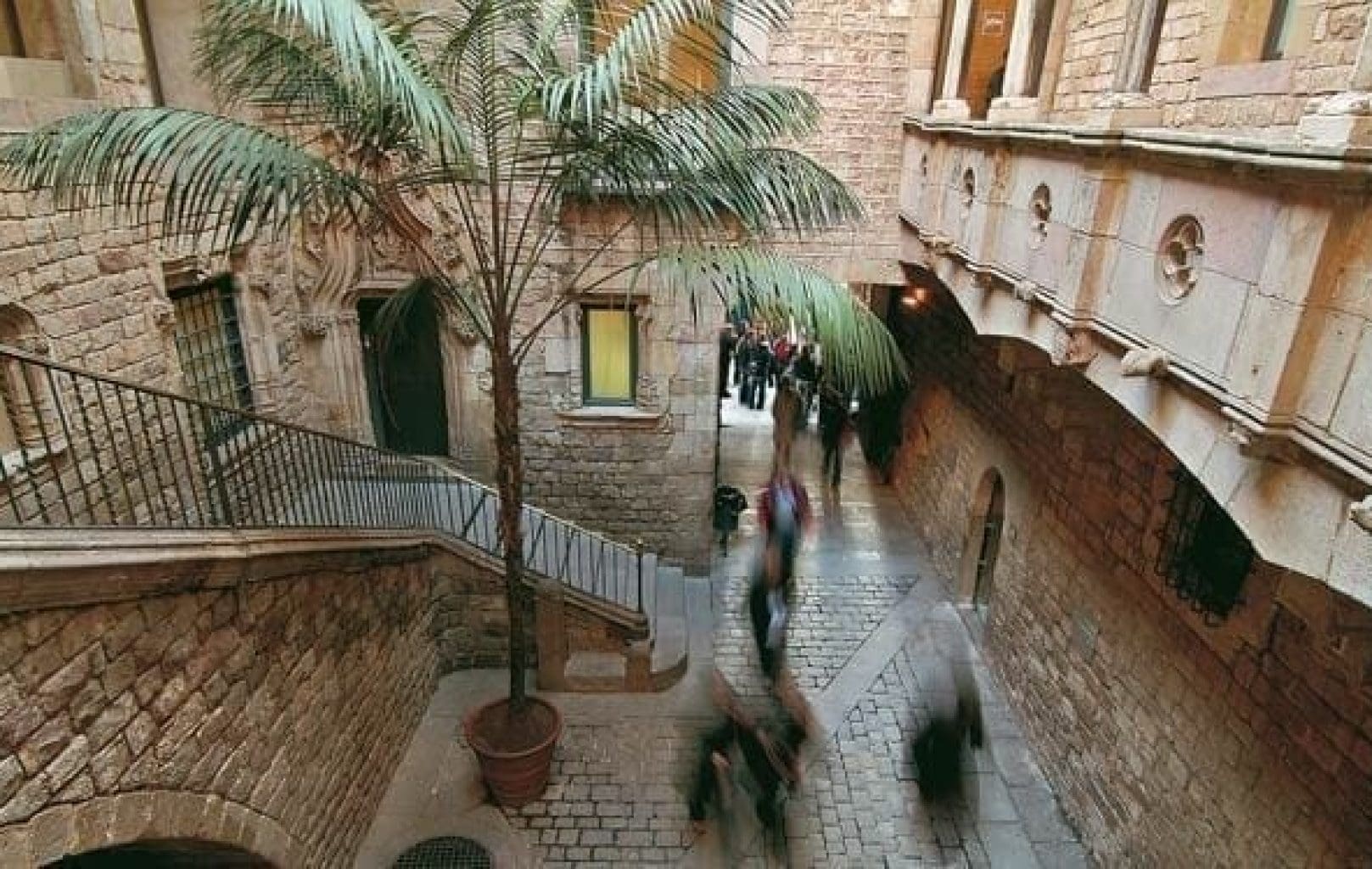 A stone courtyard near the Picasso Museum, with a large potted palm tree, arched windows, and balconies overlooking people walking on the cobblestone ground below in Barcelona.
